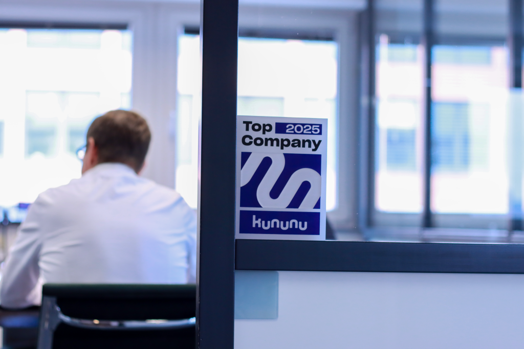 Auszeichnung für Top Company 2025 von Kununu hängt auf Glasscheibe und im Meetingraum sitzt eine Person von hinten mit weißem Hemd.